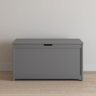 The Child Craft Harmony 33" Kids Toy Box in cool gray, featuring a flat lid and handle opening, is placed against a light-colored wall on a wooden floor.