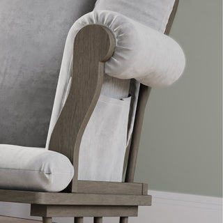 Close-up of the Child Craft Cozy Glider and Ottoman in Dapper Gray, featuring light gray cushioned fabric, wooden armrests, a small side pocket, and set against a gray wall with a white baseboard.