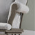 Close-up of the Child Craft Cozy Glider and Ottoman in Dapper Gray, featuring light gray cushioned fabric, wooden armrests, a small side pocket, and set against a gray wall with a white baseboard.