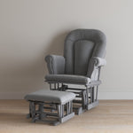 The Child Craft Cozy Glider and Ottoman in Cool Gray, with padded armrests and a matching ottoman, sits on a light wood floor against a beige wall, making it a cozy nursery chair option.