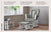 A Child Craft Tranquil Nursery Glider and Ottoman in Matte White, featuring plush foam padding, a sturdy hardwood frame, and customizable options, rests in a cozy wood-floored room beside a window, houseplant, and dollhouse.