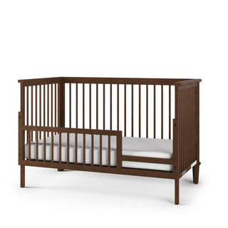 The Child Craft F09502 Toddler Guard Rail in Cocoa Bean features open slatted sides, a dark brown finish, low profile, partial guard rail on one side, and is displayed with a white mattress against a white background.