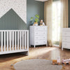 A modern nursery with a white crib, dresser, and the Child Craft F01792 Universal Changing Table Topper in Matte White on a wooden floor. A round rug, wooden toys, green accent walls, patterned wallpaper, and beige curtains complete the space.