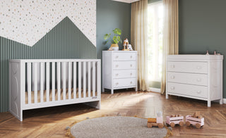 A modern nursery with a white crib, dresser, and the Child Craft F01792 Universal Changing Table Topper in Matte White on a wooden floor. A round rug, wooden toys, green accent walls, patterned wallpaper, and beige curtains complete the space.