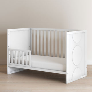 The Child Craft F09501 Toddler Guard Rail in Matte White features sturdy wood construction, a partial guard rail, and circular end panel designs. It’s shown on a light wood floor with a plain beige wall background.