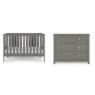 The Child Craft Atwood 3-in-1 Euro Convertible Crib in Lunar Gray with slatted sides stands beside a matching gray dresser featuring three drawers and chevron detail on the top drawer.