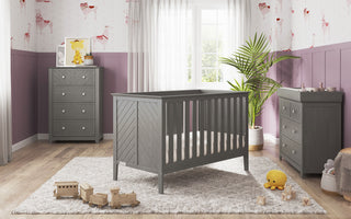 A modern nursery with a gray crib, the Child Craft Atwood 3-Drawer Dresser in Lunar Gray, and a changing table on a light rug. A wooden toy train and plush duck rest on the floor. Pink-and-white wallpaper, plant, and sunlight add warmth.