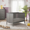 A modern nursery with gray furniture, including a crib and a Child Craft Universal Changing Table Topper in Lunar Gray. Mauve walls, toys on a light rug, and a potted plant by sheer-curtained windows complete the cozy space.