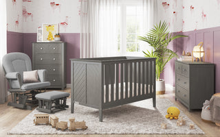 A cozy nursery features gray furniture, including the Child Craft Atwood 3-Drawer Dresser in Lunar Gray. A plush rug, toys, potted plant, and soft lighting complete the inviting space as sunlight streams through the window.