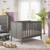 A cozy nursery with gray furniture, including a crib, rocking chair, and a Child Craft F01792 Universal Changing Table Topper in Lunar Gray on the dresser. Wooden toys, a plush duck, and plants brighten the soft rug under sheer curtain-filtered light.
