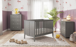 A modern nursery features the Child Craft Atwood 3-in-1 Euro Convertible Crib in Lunar Gray, a matching dresser and chest, a light rug, wooden toy train, plush toys, pastel animal wallpaper, potted plant, and sheer curtains by a bright window.