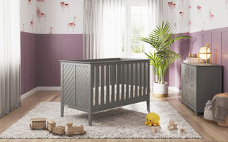 A gray crib rests on a light rug in a nursery with purple walls, wooden floors, toys, a stuffed duck, lamp, potted plant, sunny window with curtains, and the Child Craft Atwood 3-Drawer Dresser in Lunar Gray.