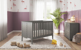 A gray wooden crib sits on a patterned rug in a nursery with purple wainscoting, ballerina wallpaper, a Child Craft Atwood 3-Drawer Dresser in Lunar Gray topped with a lamp, plus toys and plants near a window with sheer curtains.