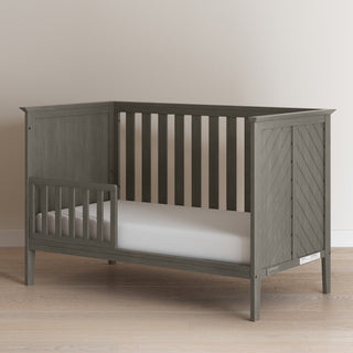 The Child Craft Atwood 3-in-1 Euro Convertible Crib in Lunar Gray features a three-sided slatted frame, chevron side panel, and white mattress, shown on a light wood floor against a neutral wall.