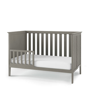 The Child Craft F09501 Toddler Guard Rail in Lunar Gray features strong wood construction, a partial guard rail, slatted sides and back, a solid mattress base, and a white mattress, all set against a plain white background.