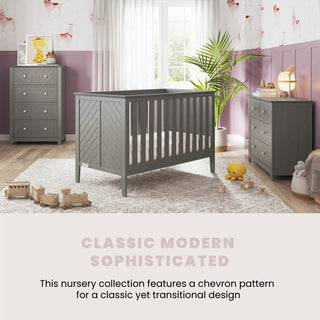 A modern nursery featuring the Child Craft Atwood 3-in-1 Euro Convertible Crib in Lunar Gray, a matching dresser and nightstand, white rug, plush toys, pink printed wallpaper, and sunlight streaming through white-curtained windows.