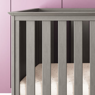 The Child Craft Atwood 3-in-1 Euro Convertible Crib in Lunar Gray sits against a purple paneled wall, featuring vertical slats and a mattress topped with a light, subtly patterned sheet.