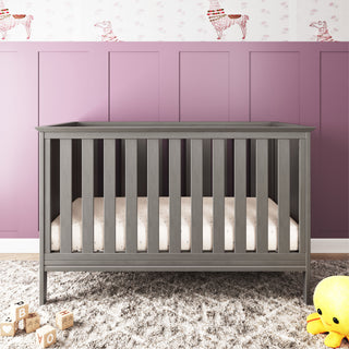The Child Craft Atwood 3-in-1 Euro Convertible Crib in Lunar Gray sits on a soft gray rug before a purple paneled wall with animal wallpaper. Wooden blocks and a yellow plush duck rest on the floor nearby.
