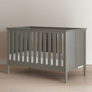 The Child Craft Atwood 3-in-1 Euro Convertible Crib in Lunar Gray features vertical slats on all sides and includes a white mattress, shown on a light wooden floor against a plain beige wall.