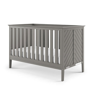 The Child Craft Atwood 3-in-1 Euro Convertible Crib in Lunar Gray features slatted sides, a chevron-patterned end panel, and comes with a white mattress inside.