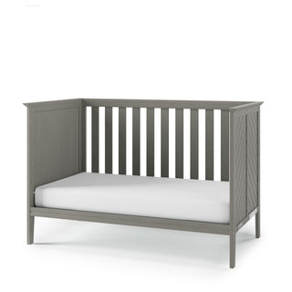 The Child Craft Atwood 3-in-1 Euro Convertible Crib in Lunar Gray features slatted sides, chevron-patterned end panels, and comes with a white mattress. Shown against a plain white background.
