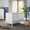 A bright, modern nursery with a white crib, matching dresser, and Child Craft F01792 Universal Changing Table Topper in Matte White; features a gray rug, wooden toys, plush orange toy, potted plant, and star-patterned blue-gray wallpaper with sunlight.