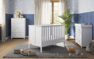 A bright, modern nursery with a white crib, matching dresser, and Child Craft F01792 Universal Changing Table Topper in Matte White; features a gray rug, wooden toys, plush orange toy, potted plant, and star-patterned blue-gray wallpaper with sunlight.