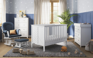 Nursery room featuring the Child Craft Atwood 3-in-1 Euro Convertible Crib in Matte White, changing table, dresser, cushioned rocking chair, star wallpaper, blue and white decor, plush rug, toys, and a potted plant by beige-curtained window.