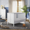 A cozy nursery with a white crib, dresser with the Child Craft F01792 Universal Changing Table Topper in matte white, blue rocking chair and ottoman, plush toys, wooden train on a gray rug, star-patterned blue and white walls, and beige-curtained window.