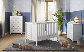 A nursery features the Child Craft Atwood 3-in-1 Euro Convertible Crib in matte white with a matching dresser and chest, a gray rug, wooden toys, plush bear, lamp, blue star wallpaper, potted plant, and sunlight streaming in.