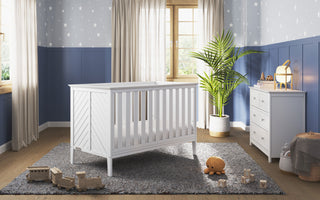 A cozy nursery featuring the Child Craft Atwood 3-in-1 Euro Convertible Crib in Matte White, plush and wooden toys, gray rug, white dresser, large potted plant, star-patterned wallpaper, and two windows with beige curtains letting in sunlight.