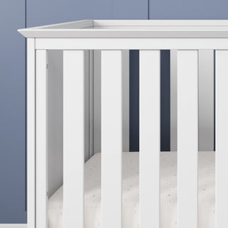 A close-up of the Child Craft Atwood 3-in-1 Euro Convertible Crib in Matte White, featuring vertical slats and a subtly patterned mattress sheet, set against a blue-paneled wall.