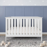 The Child Craft Atwood 3-in-1 Euro Convertible Crib in Matte White stands on a gray textured rug with wooden alphabet blocks nearby. Behind it is a blue paneled wall with a white star border.