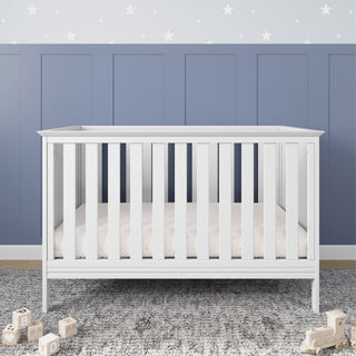 The Child Craft Atwood 3-in-1 Euro Convertible Crib in Matte White stands on a gray textured rug with wooden alphabet blocks nearby. Behind it is a blue paneled wall with a white star border.