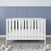 The Child Craft Atwood 3-in-1 Euro Convertible Crib in Matte White stands on a gray textured rug with wooden alphabet blocks nearby. Behind it is a blue paneled wall with a white star border.
