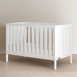 The Child Craft Atwood 3-in-1 Euro Convertible Crib in Matte White, featuring slatted sides and angled panels, is displayed on a light wood floor against a beige wall.