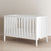 The Child Craft Atwood 3-in-1 Euro Convertible Crib in Matte White, featuring slatted sides and angled panels, is displayed on a light wood floor against a beige wall.