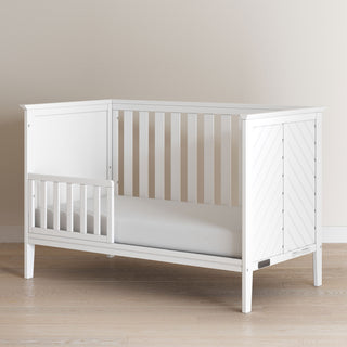 The Child Craft Atwood 3-in-1 Euro Convertible Crib in Matte White features slatted sides and a partial guardrail, shown in a room with light walls and wood flooring.