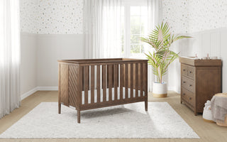A modern nursery features a wooden crib, a Child Craft Atwood 3-Drawer Dresser in Cocoa Bean, a potted plant, and soft natural light through sheer curtains. Light walls with subtle polka dots complete the look.