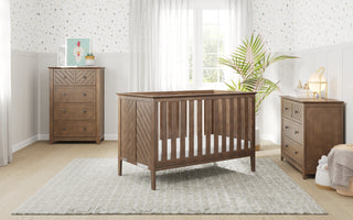 A cozy nursery features a wooden crib, two Child Craft Atwood 3-Drawer Dressers in Cocoa Bean, a white rug, soft patterned wallpaper, and a potted plant by a sunlit window with sheer curtains.