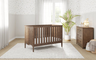 A wooden baby crib sits on a white rug in a bright nursery with the Child Craft Atwood 3-Drawer Dresser in Cocoa Bean, a potted plant, basket, and polka dot wallpaper. Sunlight streams through sheer curtains over tall windows.