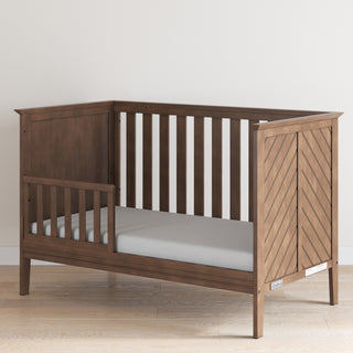 The Child Craft F09501 Toddler Guard Rail in Cocoa Bean is shown with a chevron-patterned end panel and light gray mattress, set on a light wood floor—ideal for an easy crib-to-toddler bed conversion.