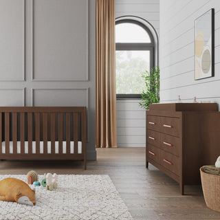 A modern nursery features a wooden crib, Child Craft's Kieran 6-Drawer Double Dresser in Toasted Chestnut, and a large window with tan curtains. Toys and a stuffed fox rest on a rug; a teddy bear sits in a basket against shiplap and gray paneled walls.