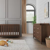 A modern nursery with a wooden crib, matching dresser featuring the Child Craft F01792 Universal Dressing Kit in Toasted Chestnut, a white textured rug with toys, stuffed fox and teddy bear in a basket, gold curtains, and framed abstract art.
