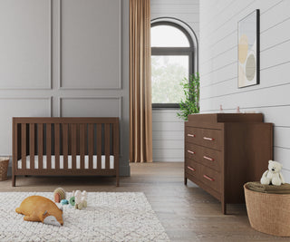 A modern nursery with a wooden crib, matching dresser featuring the Child Craft F01792 Universal Dressing Kit in Toasted Chestnut, a white textured rug with toys, stuffed fox and teddy bear in a basket, gold curtains, and framed abstract art.