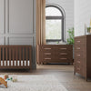 A modern nursery with a wooden crib, Child Craft Kieran 6-Drawer Double Dresser in Toasted Chestnut, chest of drawers, beige curtains on the window, plush rug, fox toy, plant décor, and framed art for a minimalist look.