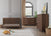 A modern nursery with wooden furniture, including a crib, a dresser, and the Child Craft Kieran 5-Drawer Chest in Toasted Chestnut. A plush fox toy and balls rest on a textured rug, with sunlight streaming through an arched window with beige curtains.