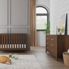 A modern nursery with a wooden crib, a Child Craft Kieran 6-Drawer Double Dresser in Toasted Chestnut, gold curtains, a soft rug with toys and a pillow, plus plants and a framed picture decorating the dresser.