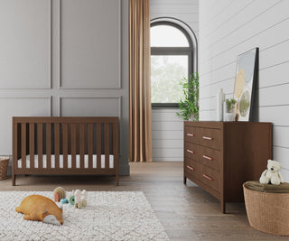 A modern nursery with a wooden crib, a Child Craft Kieran 6-Drawer Double Dresser in Toasted Chestnut, gold curtains, a soft rug with toys and a pillow, plus plants and a framed picture decorating the dresser.