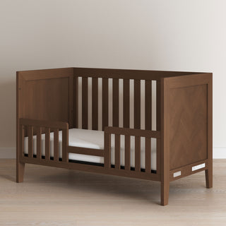 The Child Craft Kieran 3-in-1 Convertible Crib in Toasted Chestnut features open side slats and a partial guardrail, shown on a light wood floor against a plain, light-colored wall.